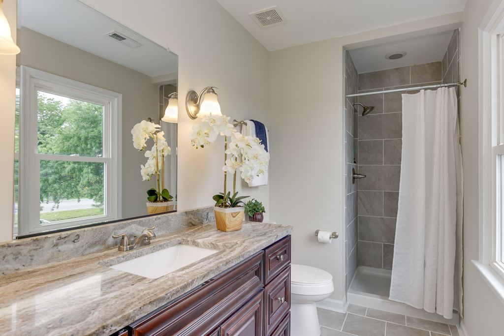 Bathroom with a marble countertop, dark wooden cabinets, a large mirror, shower with gray tiles, and white curtain.