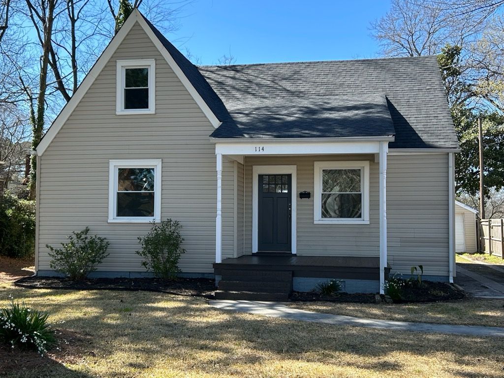 A light-colored, two-story house with a dark roof, small front porch, and manicured lawn. The house has a door and windows.