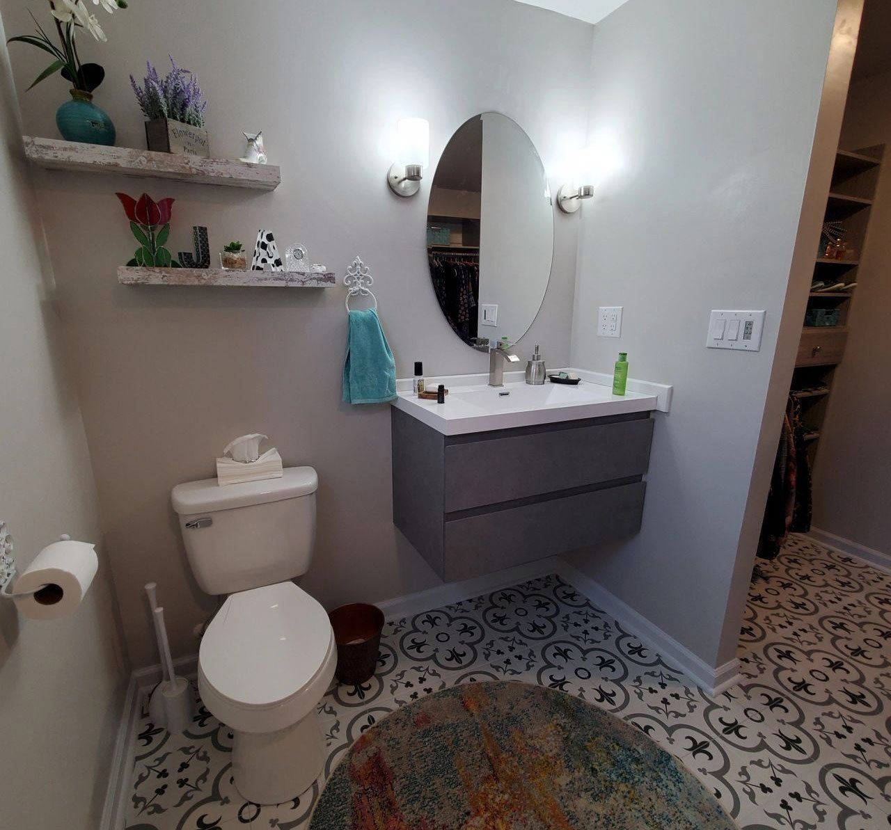 Bathroom with a grey floating vanity, oval mirror, and patterned floor. A toilet is on the left and a rug in the foreground.