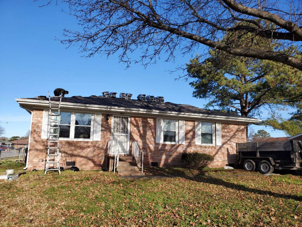 A brick house with a damaged roof under a clear blue sky, ladder leaning against the side.