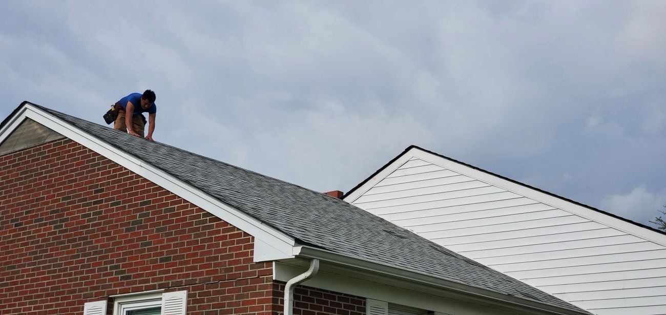 A person is working on the roof of a brick house. They are silhouetted against a cloudy sky.