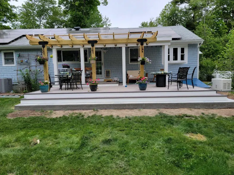 Backyard deck with pergola; blue house, chairs, plants.