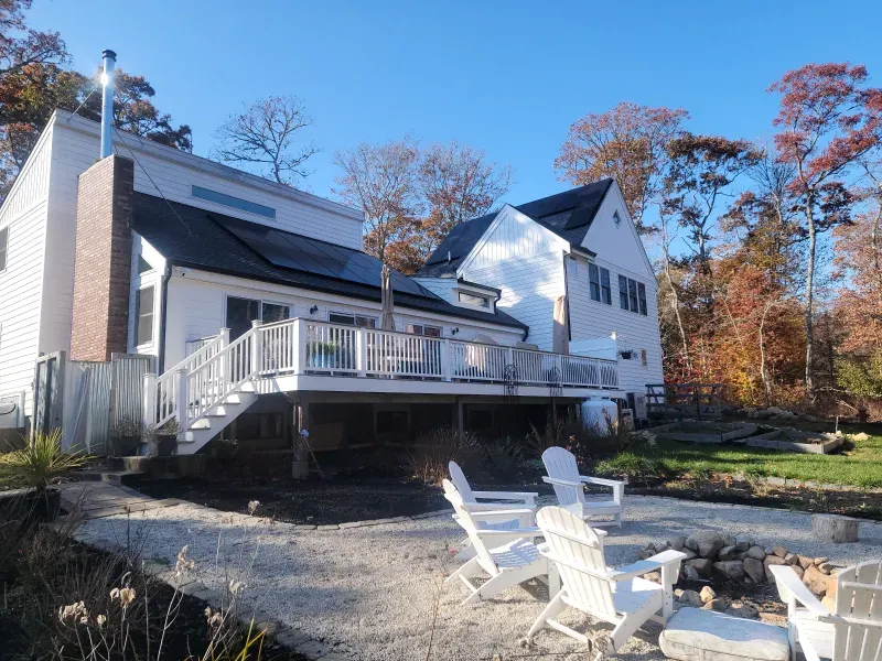 A person digs a trench in a backyard near a small excavator, a tree, and a house.