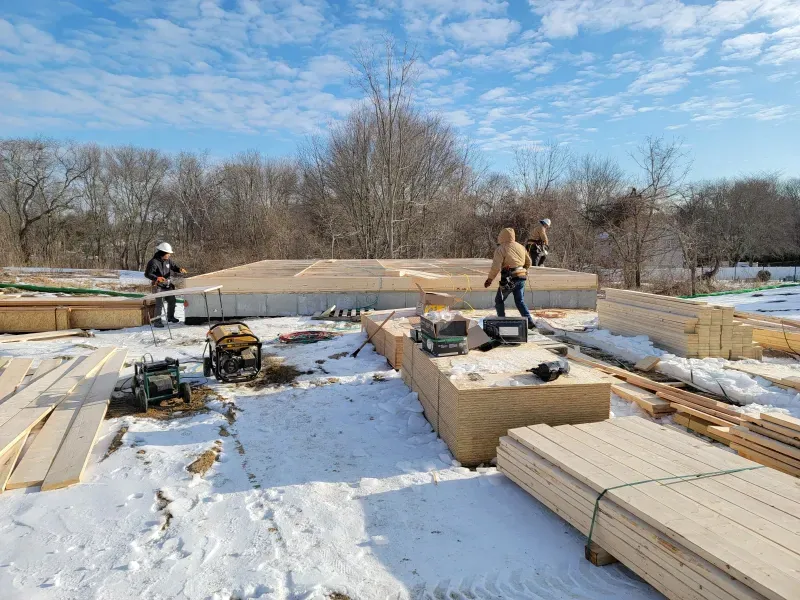 A person digs a trench in a backyard near a small excavator, a tree, and a house.