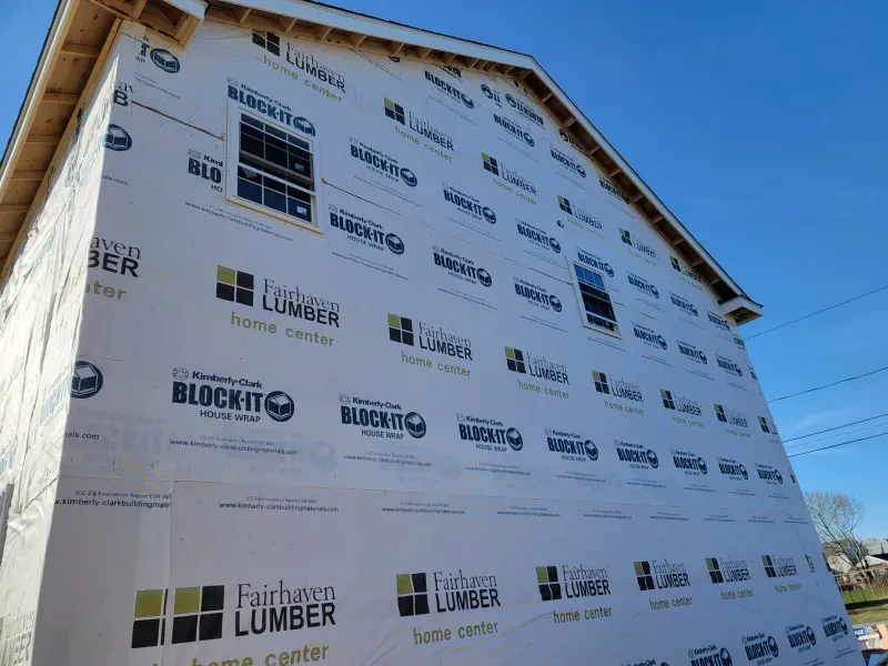 Exterior of a house under construction; white weather-resistant barrier, partially framed with window openings.