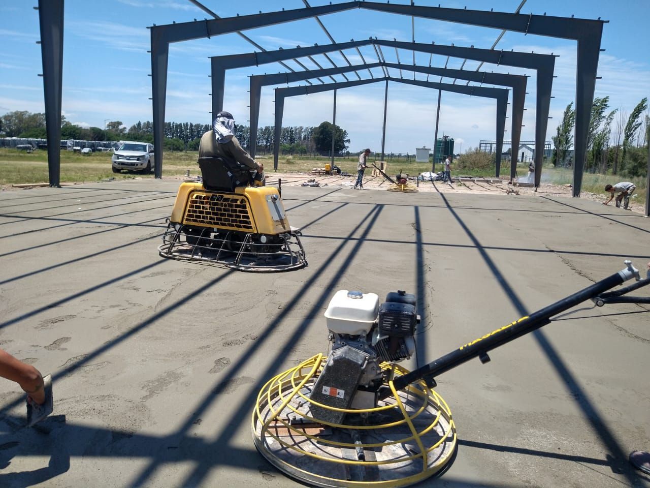 Trabajadores alisan el piso de concreto húmedo debajo de un edificio con estructura de acero en un día soleado.