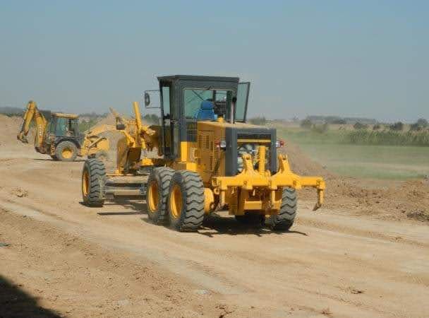 Una motoniveladora amarilla allana un camino de tierra; una excavadora al fondo. Un día soleado y brillante.