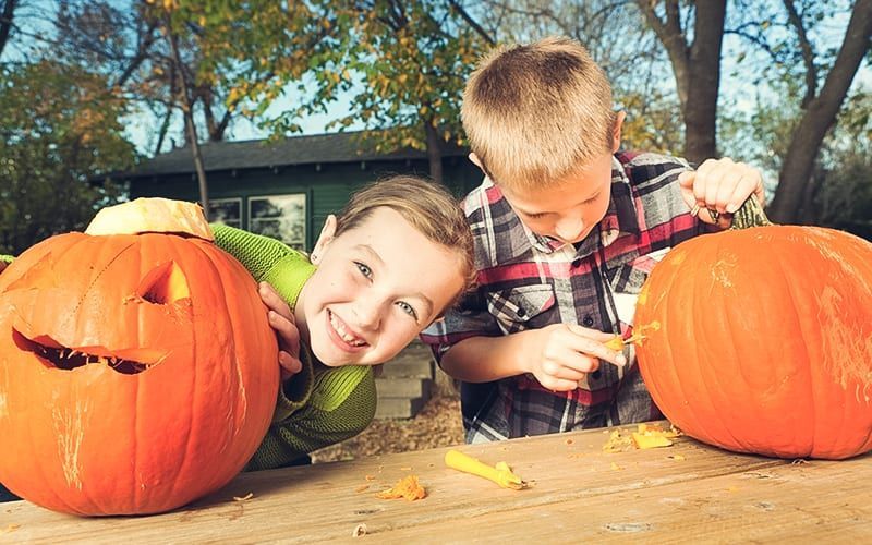 Two kids carving pumpkins outside on a sunny day. One smiles, the other carves with a tool.