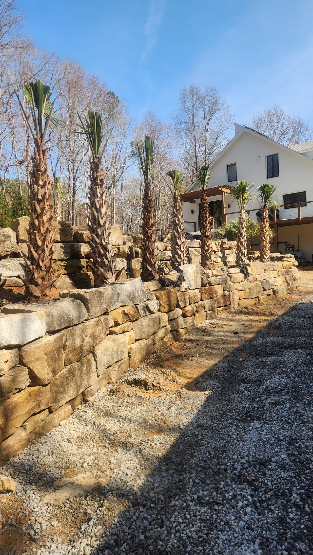 Stone retaining wall with leafless vines in a yard, gravel path, and a white house in the background