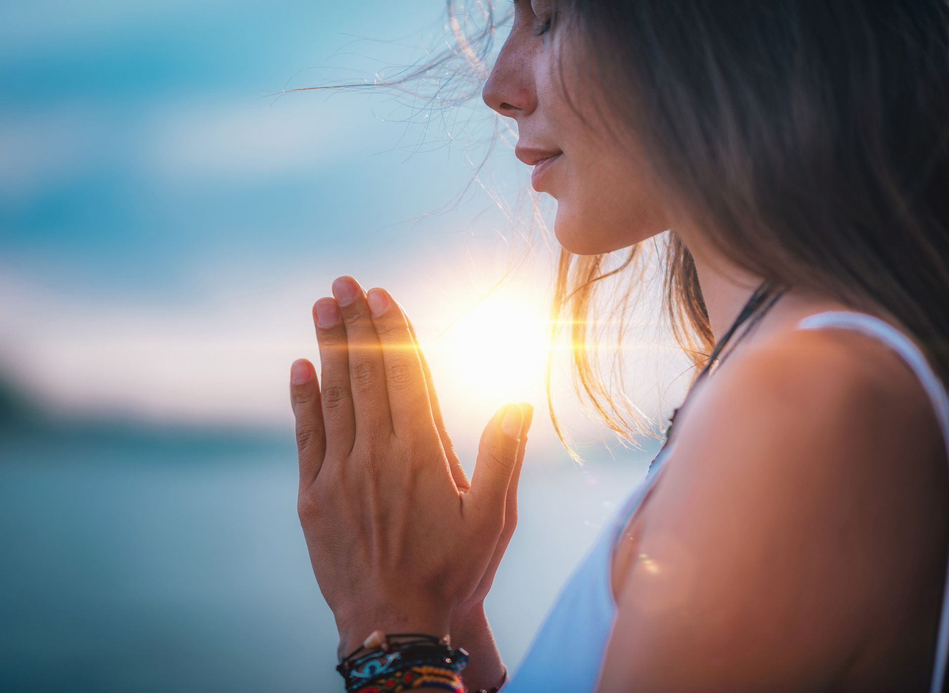 Woman in white top with hands together, eyes closed, bathed in sunlight.