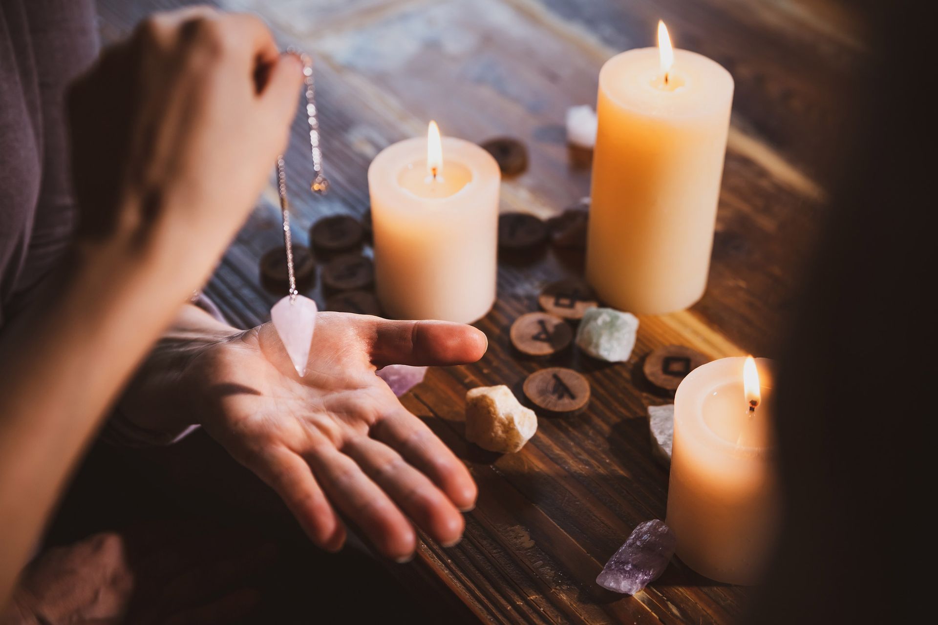 A person uses a pendulum above an open palm surrounded by candles, crystals, and rune stones.