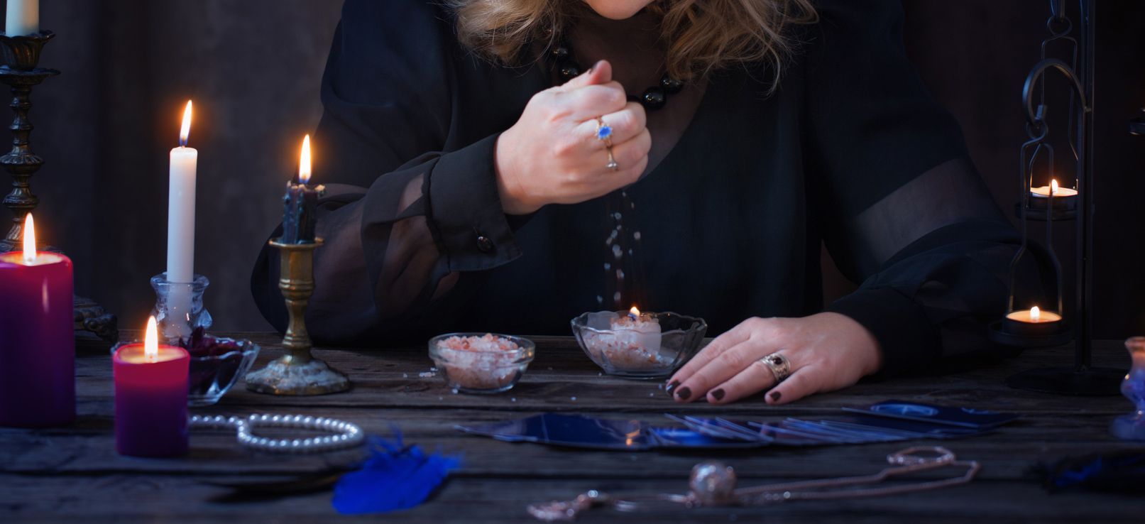 A person with rings, dressed in black, sprinkles something into a bowl. Candles and crystals are on a dark table.