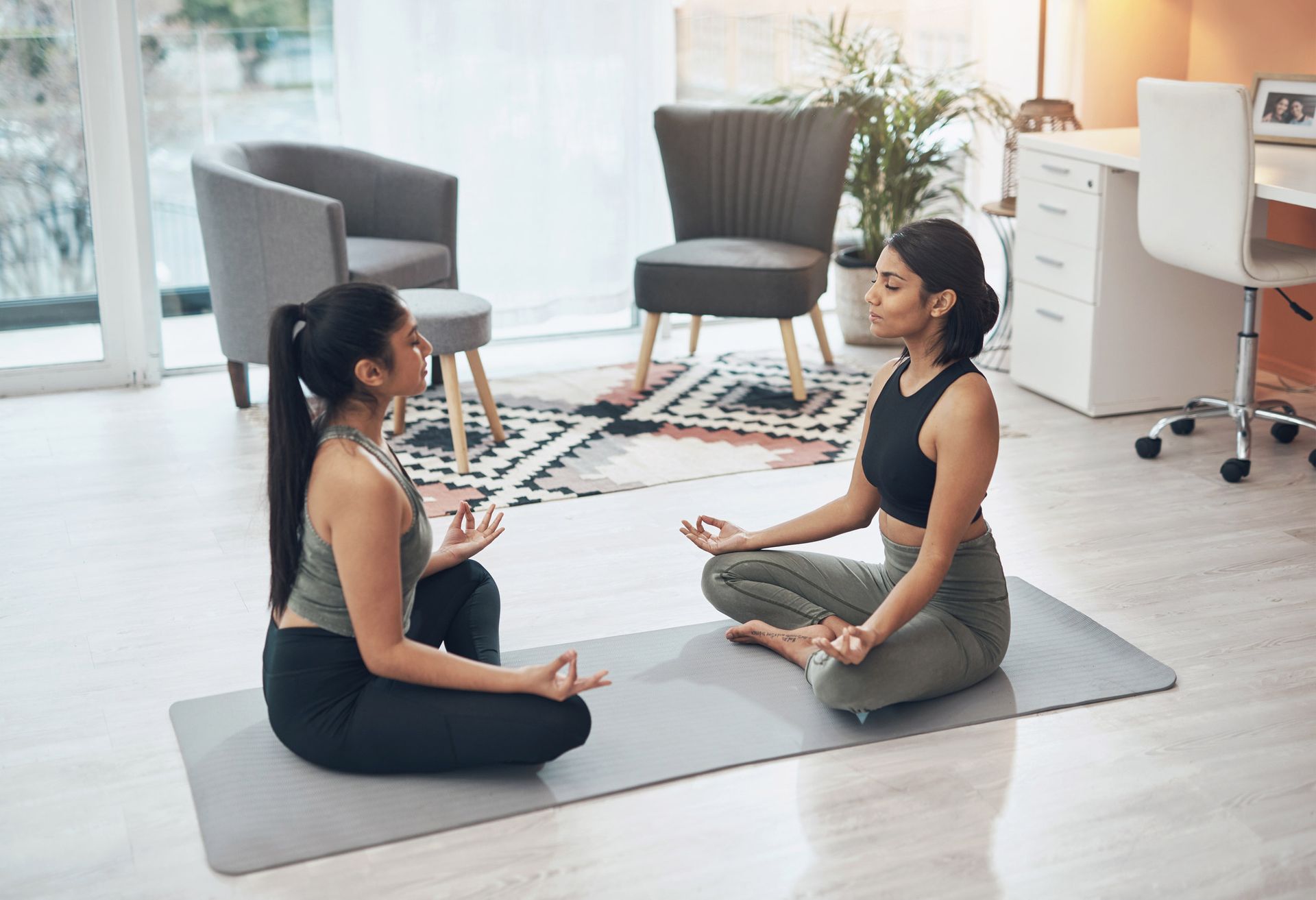 Two women meditate on a yoga mat indoors, hands in prayer position.