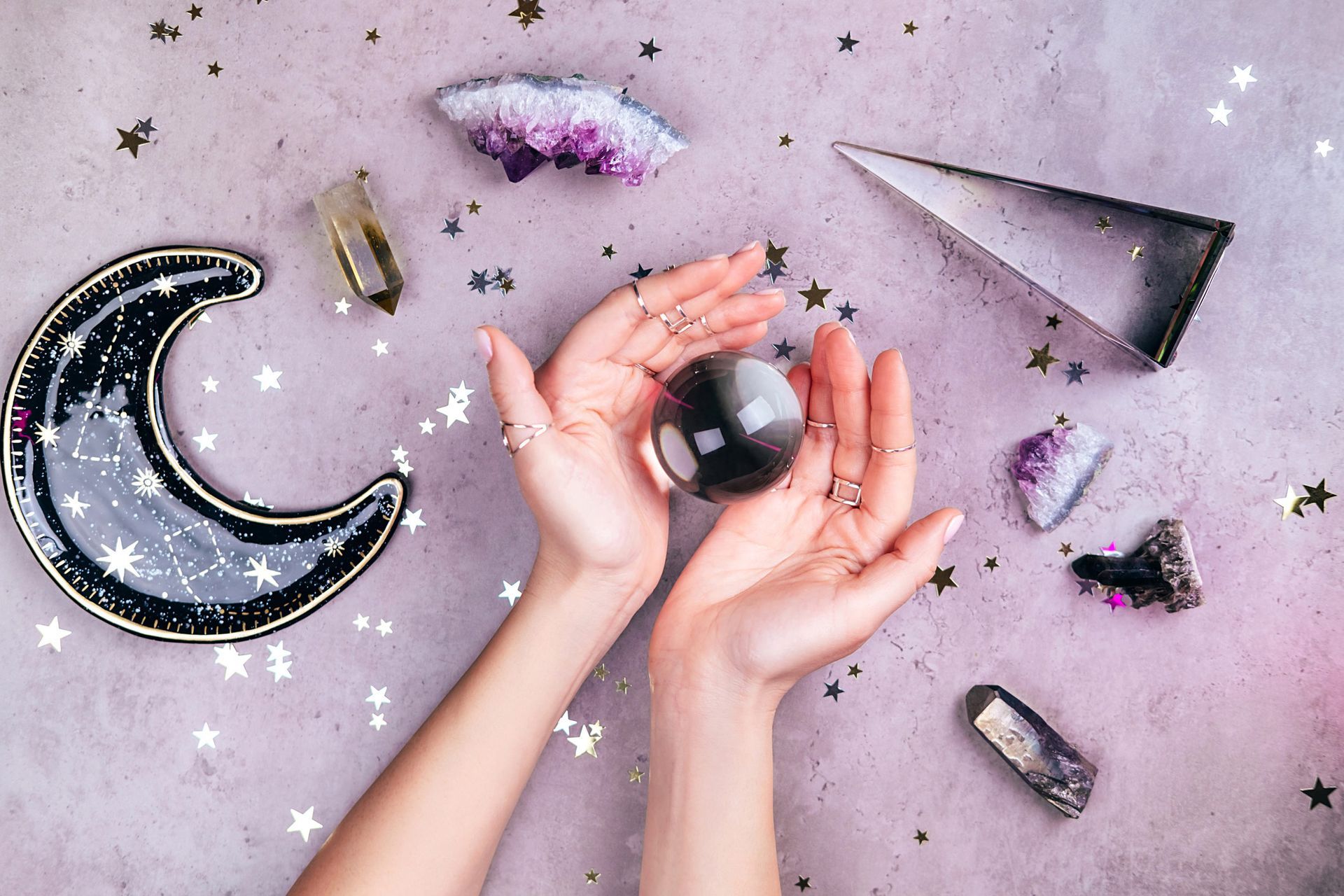 Hands holding a crystal ball amidst crystals, a moon-shaped dish, and stars on a purple surface.