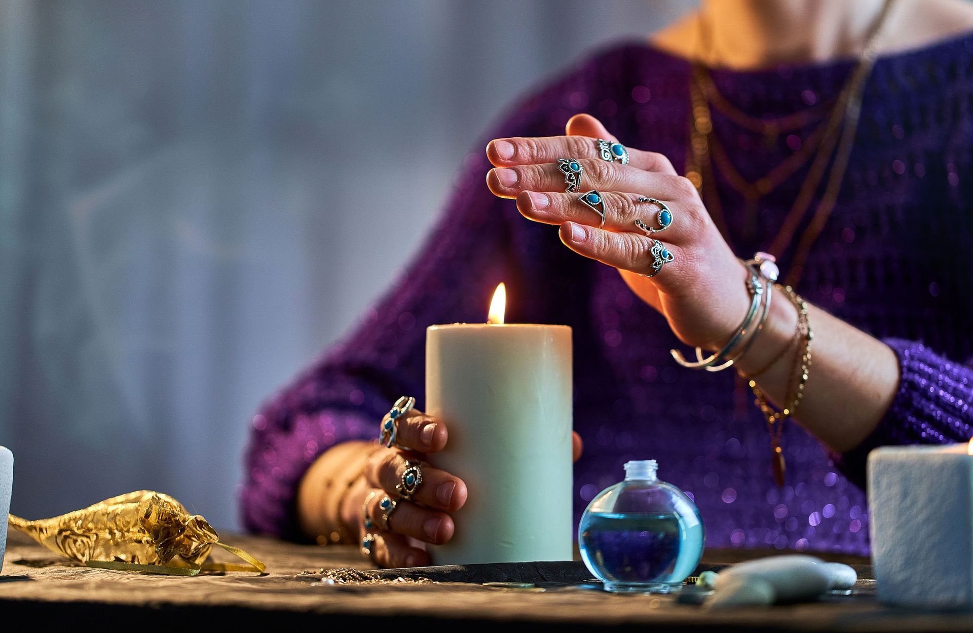 Person with jewelry, hands over a lit candle, dark setting.
