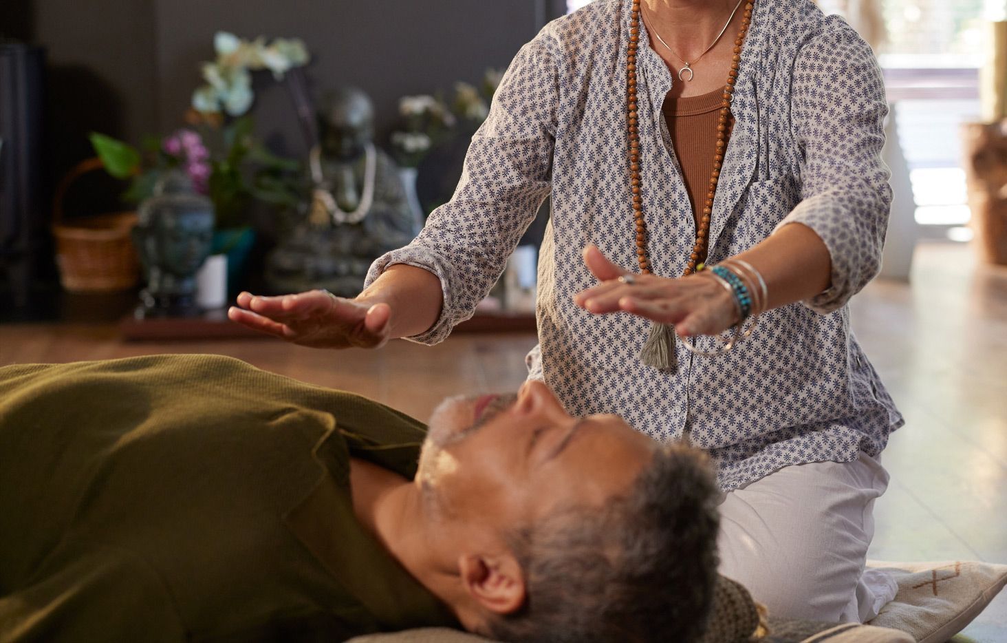Woman performing Reiki on a man lying down. Hands are over his chest. Indoors, near a buddha statue.