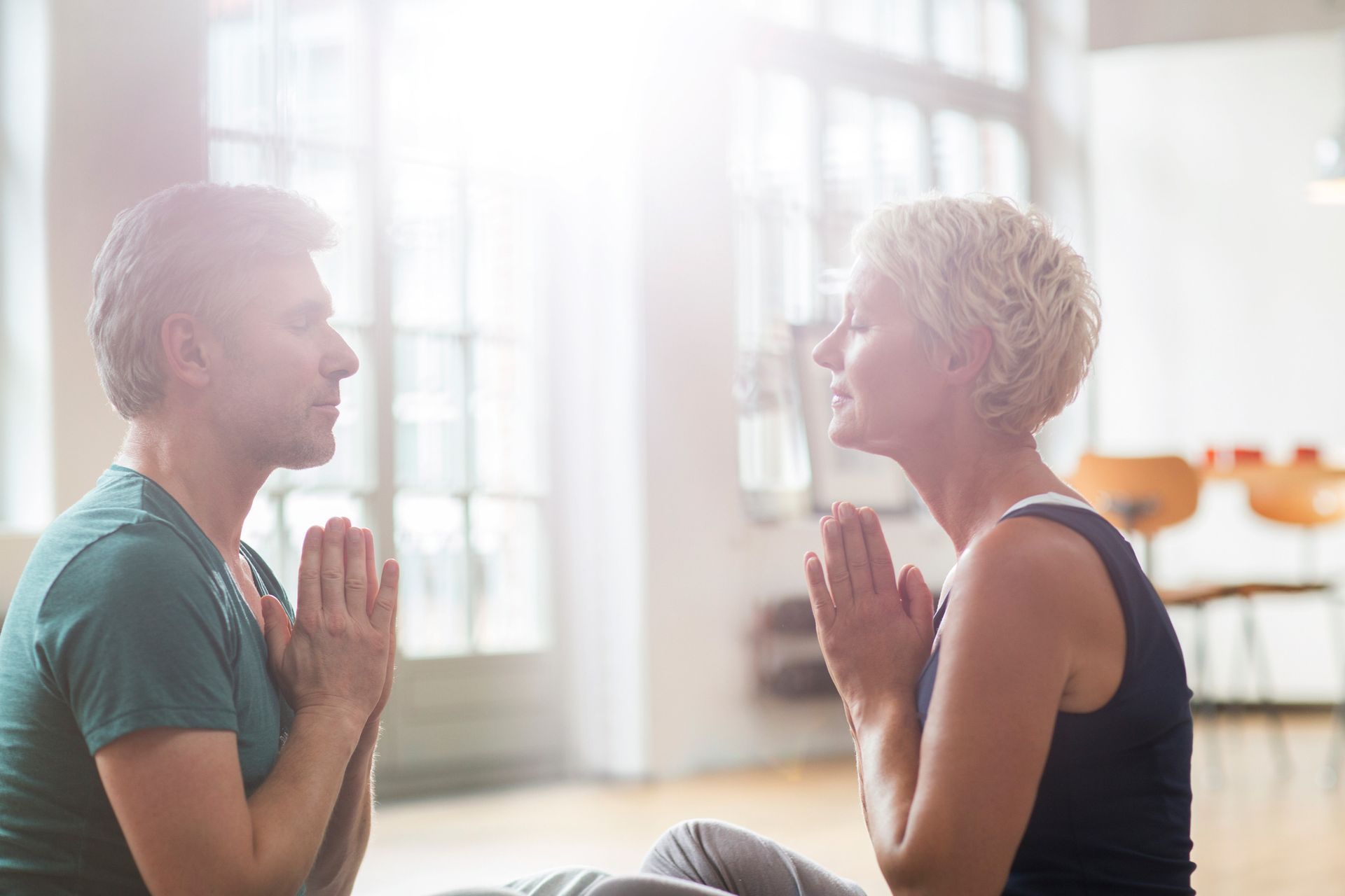 Two people, facing each other with hands together in prayer pose, meditating in a bright room.