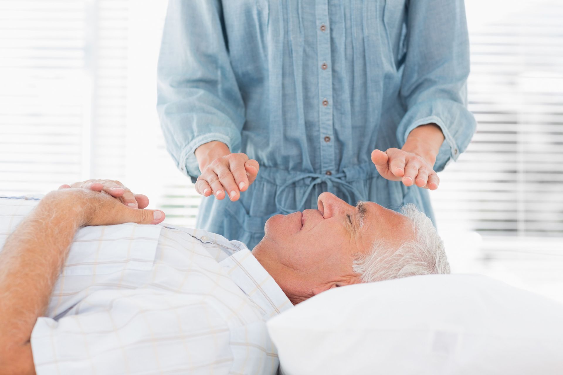 Older man receiving hands-on healing therapy; woman hovers hands over his head in a light-filled room.