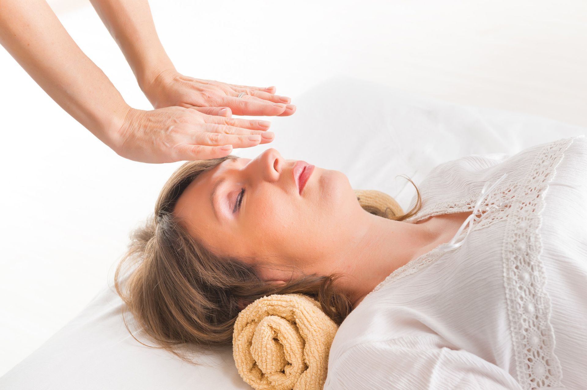 Woman receiving reiki treatment, hands hovering over her head. White room, peaceful expression.