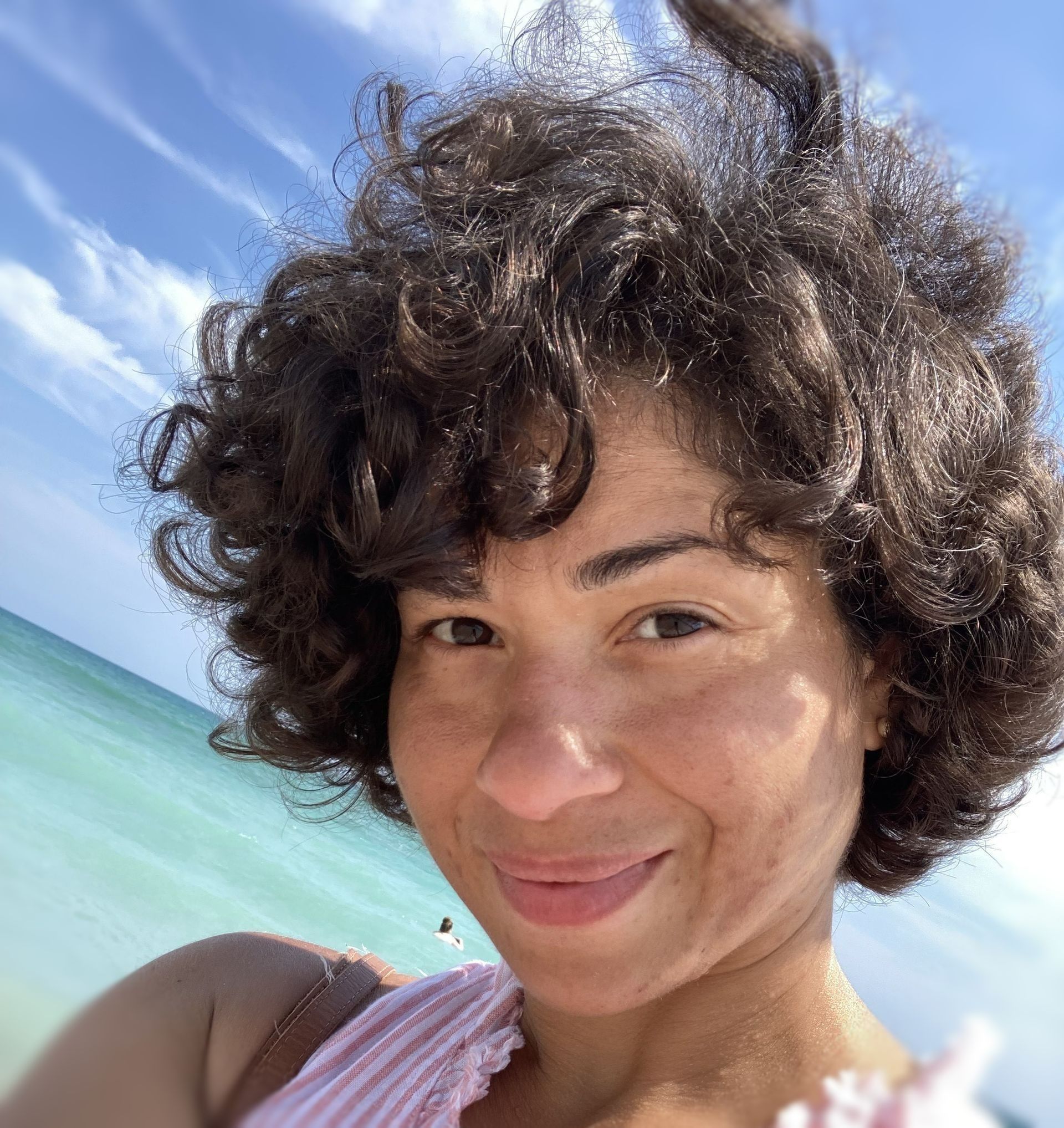 Woman with curly dark hair smiles on a beach under blue sky; turquoise water visible.