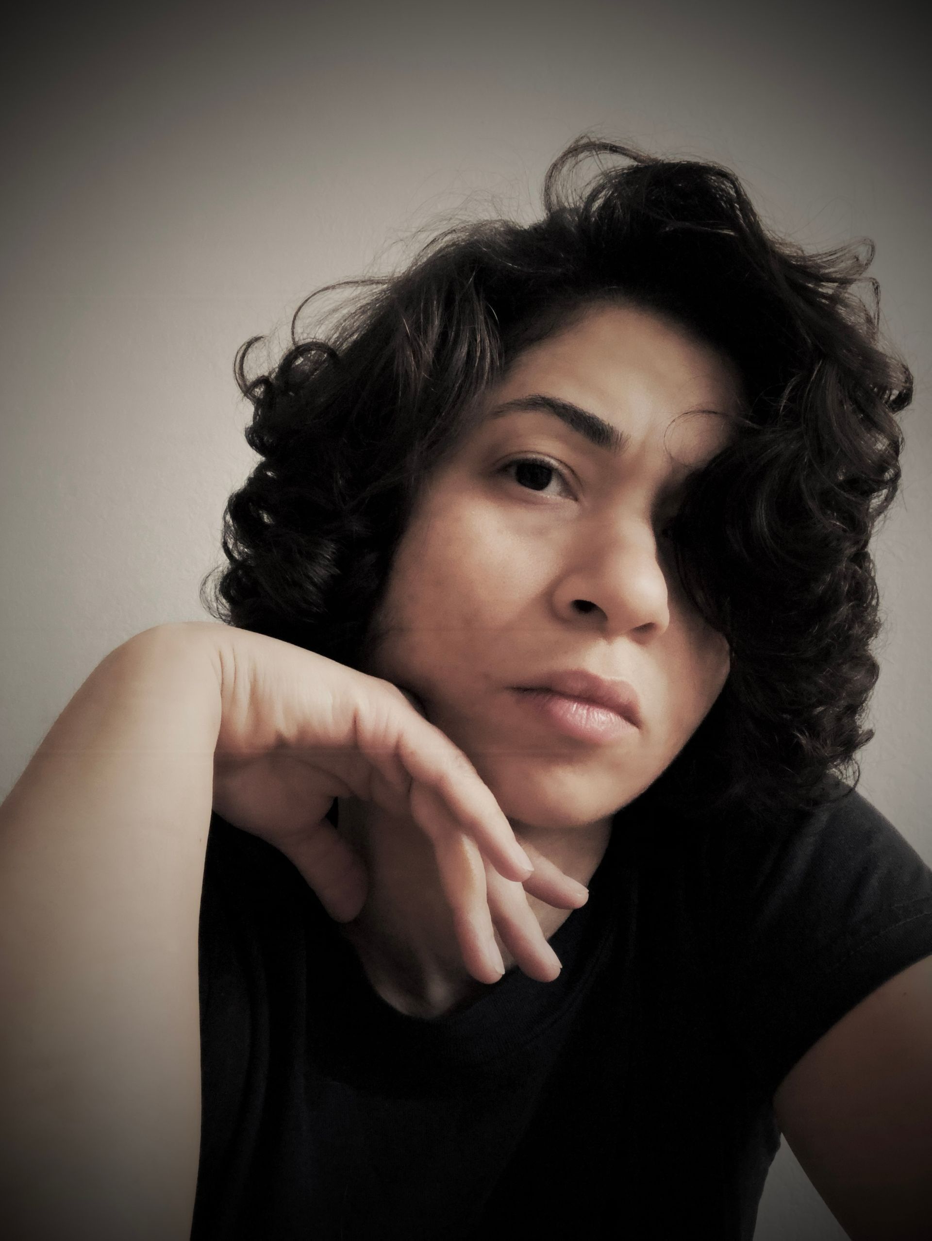 Woman with dark curly hair, resting chin on hand, looking directly at the viewer.