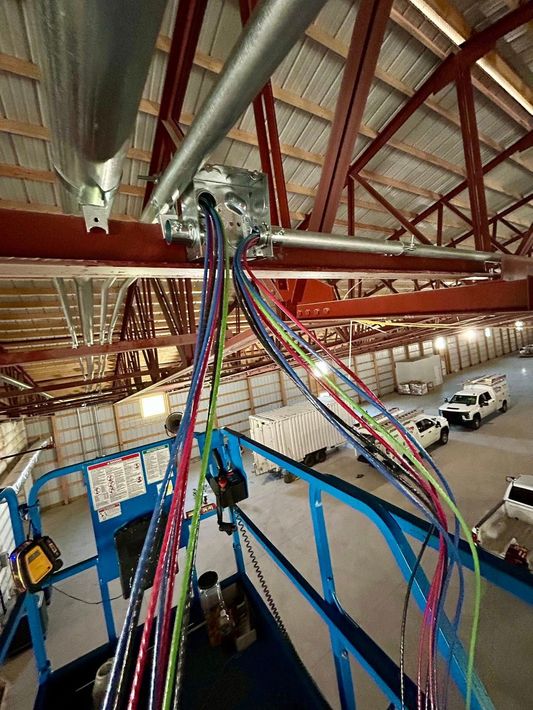 Electrical wires hang from a metal junction box on a ceiling beam above a blue aerial work platform in a large warehouse.