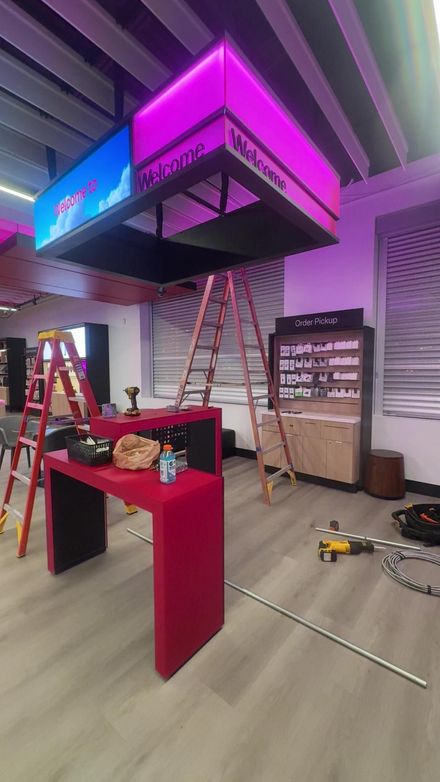 A bright pink reception desk and ladders are set up under a glowing, suspended sign in a modern, commercial interior.