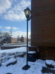 A tall, black lamppost stands on a snowy patch of ground next to a red brick building under a sunny, partly cloudy sky.