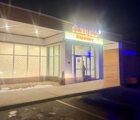 A night shot of a Sunnyside Dispensary storefront with bright, lit signage and a glass entrance door.