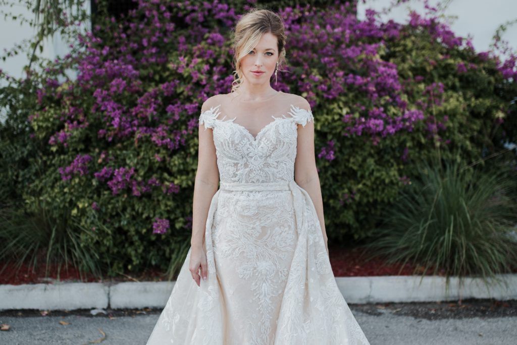 Bride in a beaded wedding dress with off-the-shoulder straps, standing outdoors with a purple flower bush.