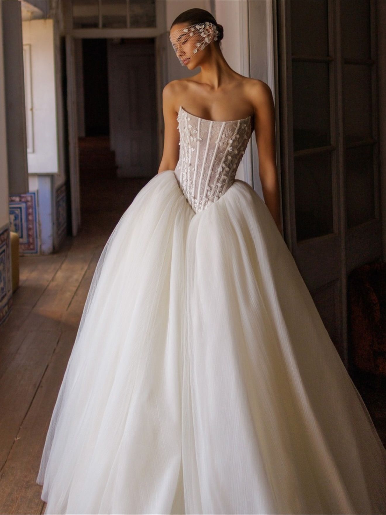 Woman in a white strapless ball gown, wearing a delicate headpiece, standing indoors.