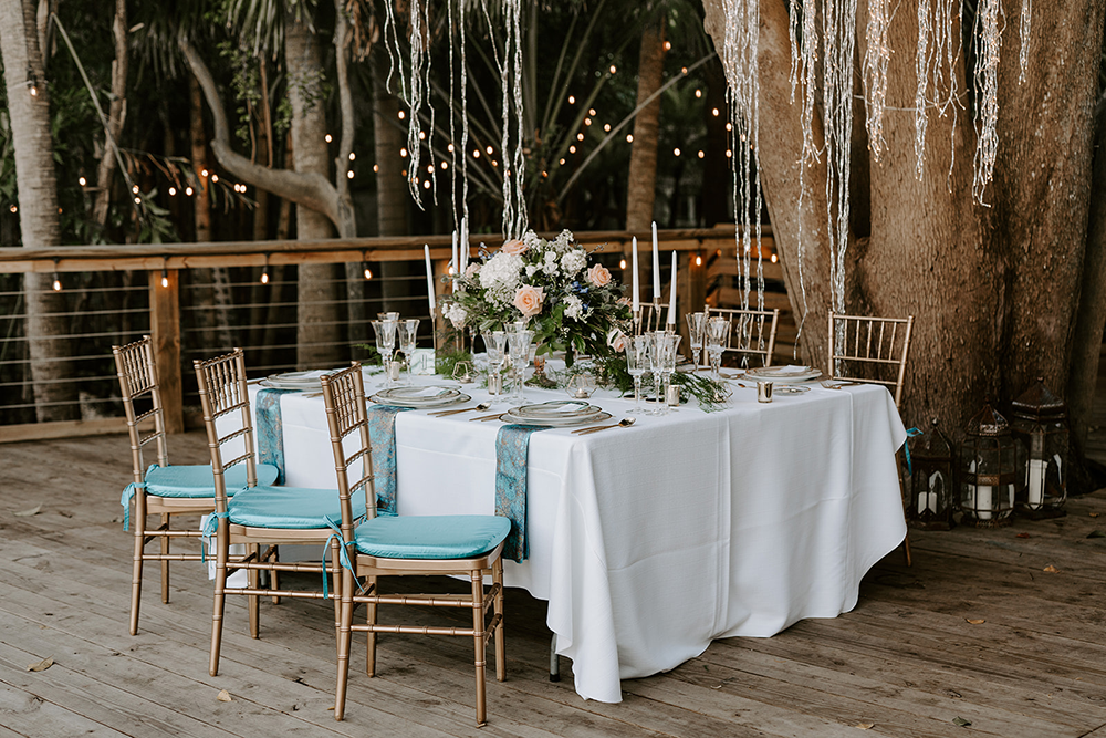 Table set for an outdoor event with white tablecloth, gold chairs, blue accents, and floral centerpiece.