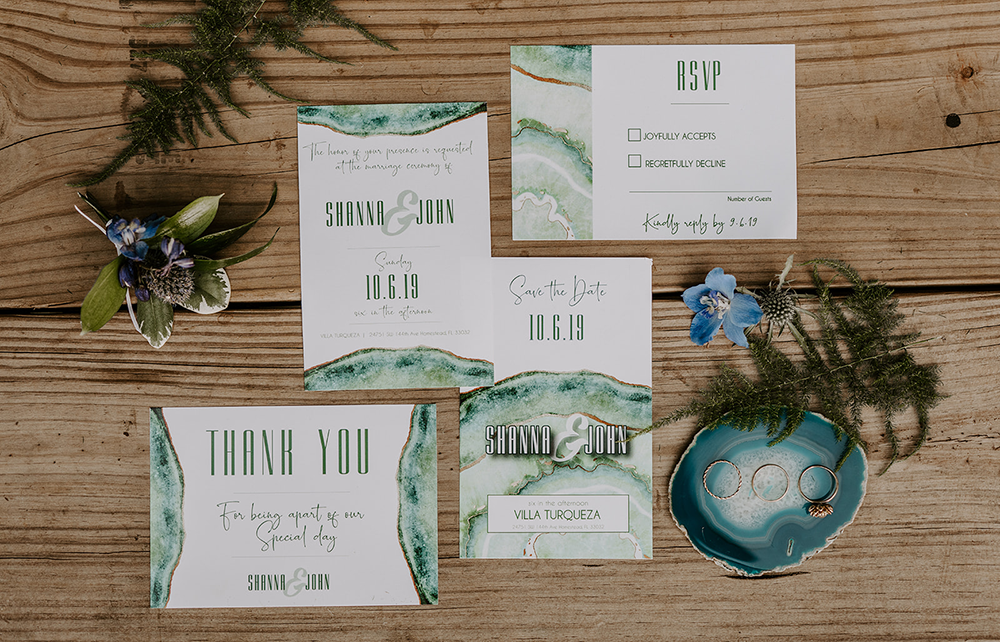 Wedding invitation suite on wood: watercolor green accents, boutonniere, ring holder, and thank you card.