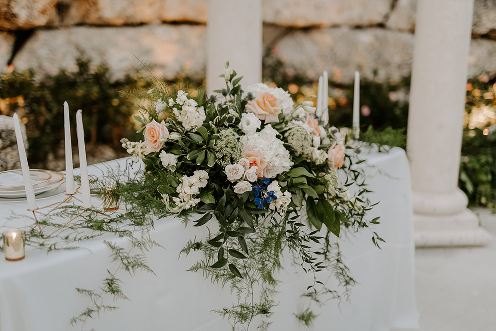Wedding table with floral centerpiece and candles, near stone pillars and wall.