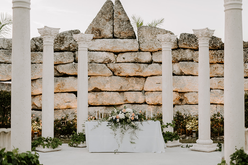 Wedding altar with white pillars, stone wall backdrop, floral arrangement, and candles.