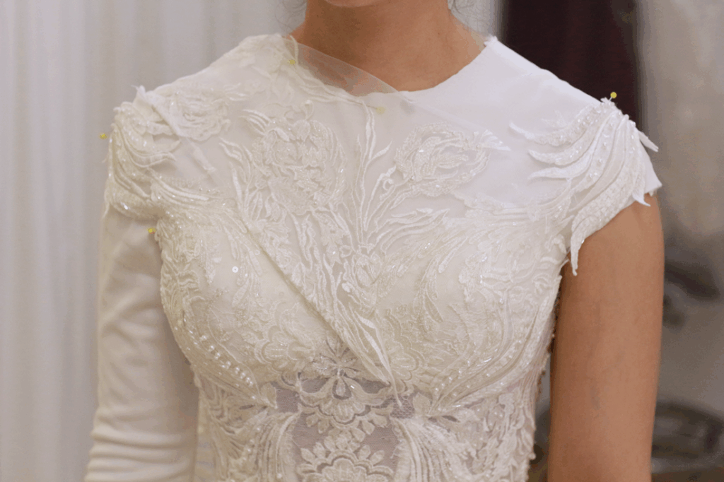 Woman wearing a white wedding dress with lace detail, one long sleeve, being fitted.