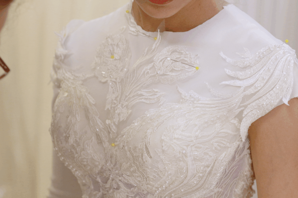 Woman in white dress being fitted, covered in floral lace, pins.