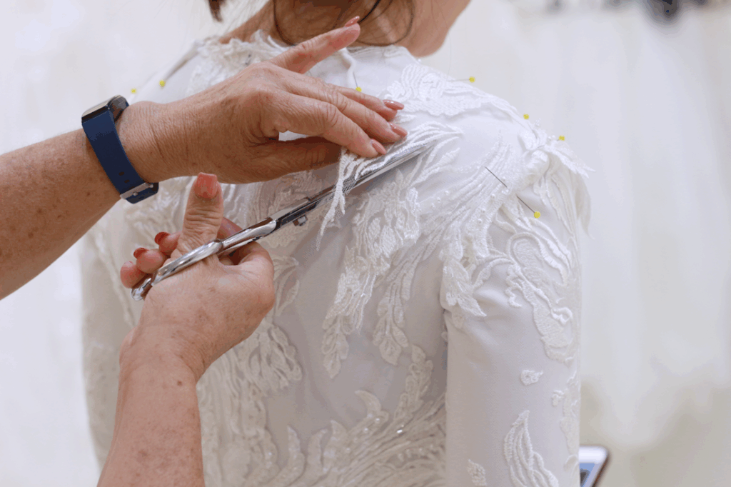 Person trimming fabric on the back of a white, embellished dress. White background.