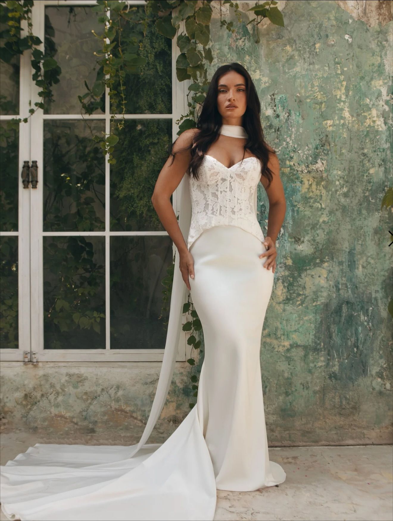 Woman in white wedding dress with cape, standing by a window with vines.