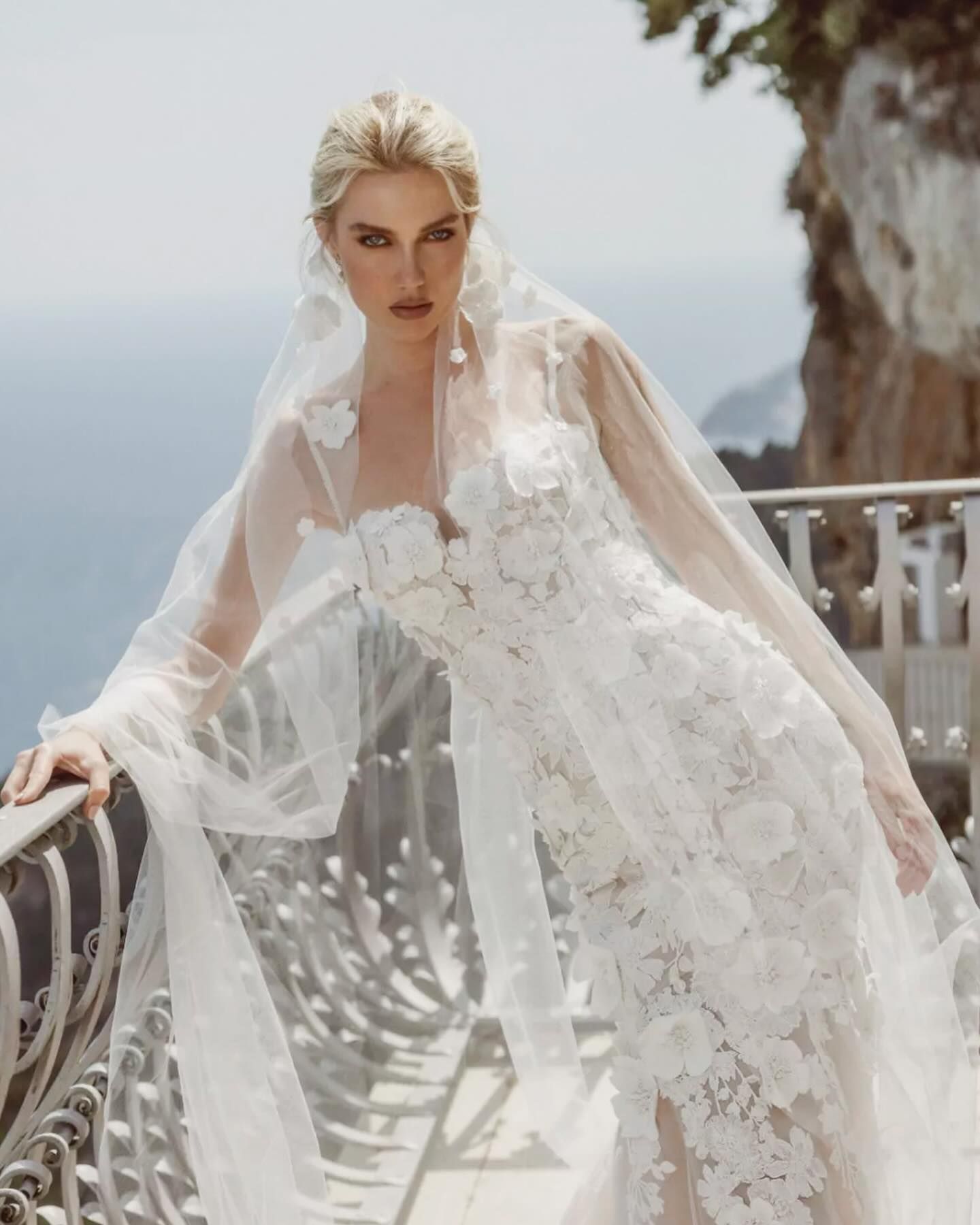 Bride in white floral wedding dress with veil, leaning on a balcony railing with a scenic backdrop.