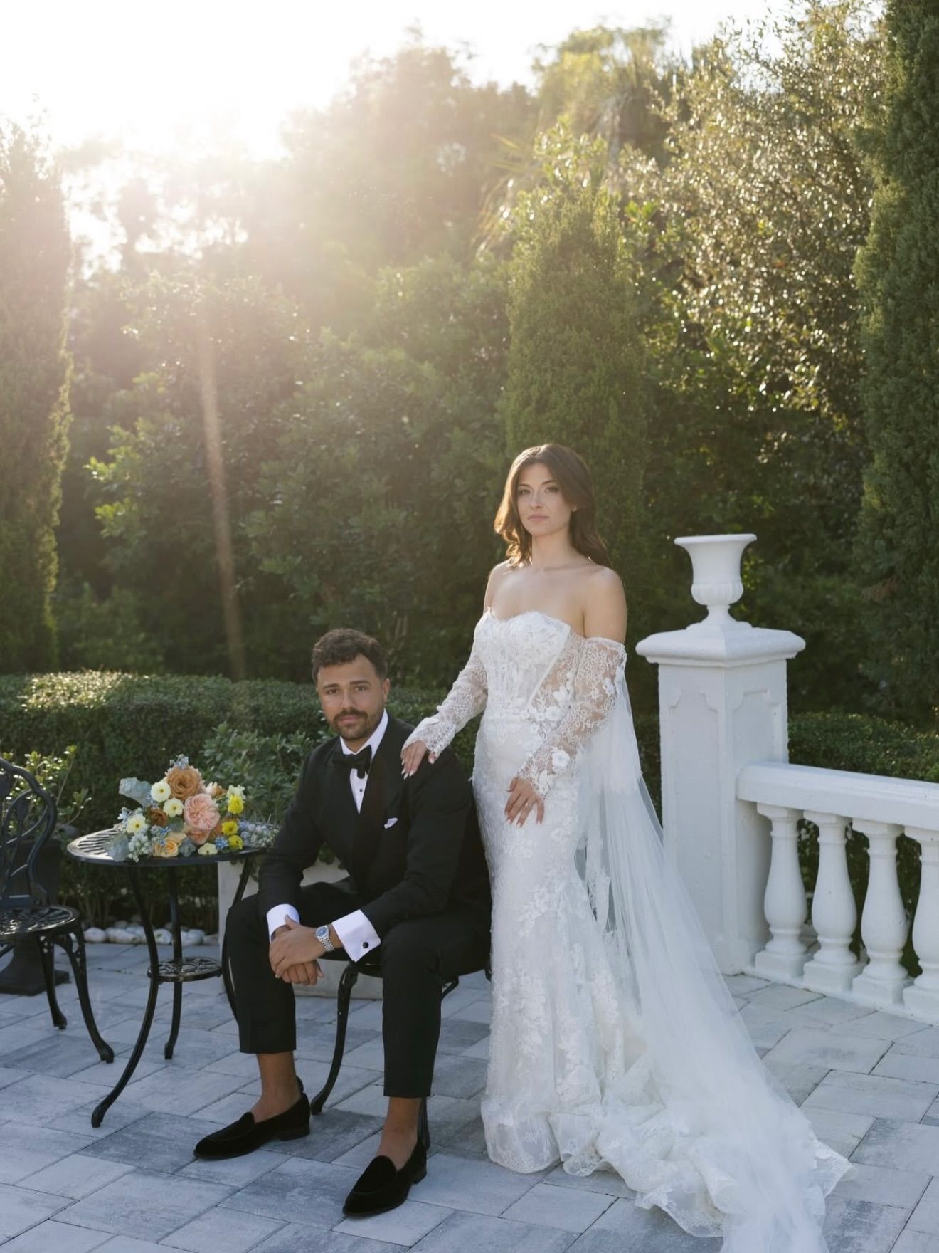 Couple poses outdoors, man in black suit, woman in white gown with veil.