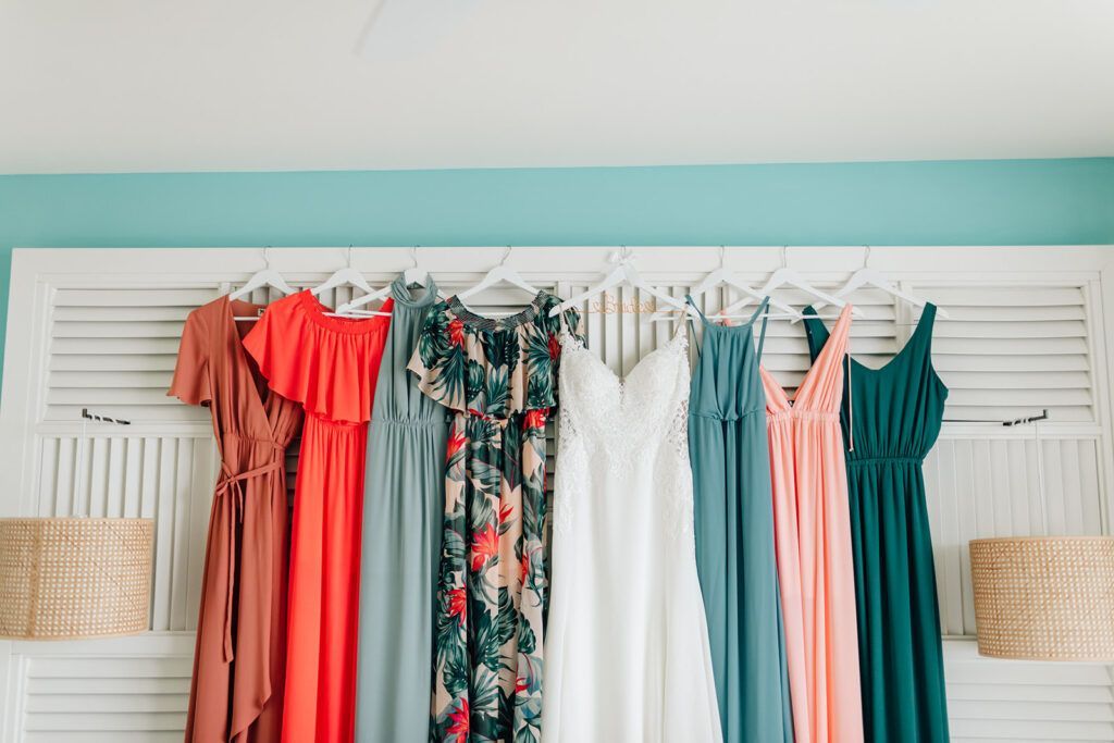 Bridesmaid dresses in various colors hanging on a white shuttered headboard against a teal wall.