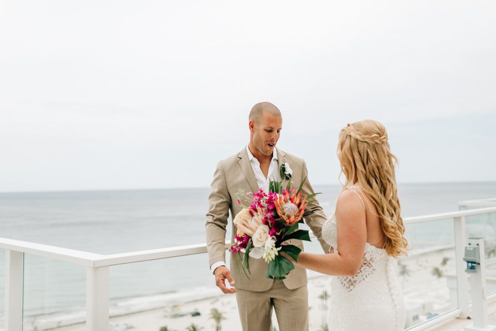 Groom in tan suit surprised by bride in white dress, ocean view, holding bouquet.