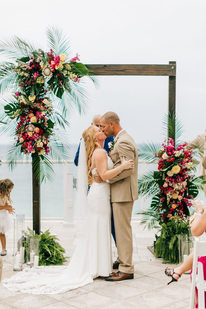 Couple kissing under a floral arch during an outdoor wedding ceremony.