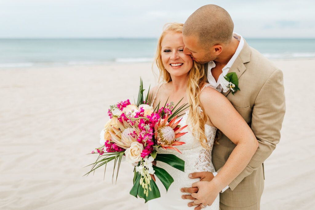 Couple on beach; man kissing woman's cheek, holding her waist, she holds a flower bouquet.