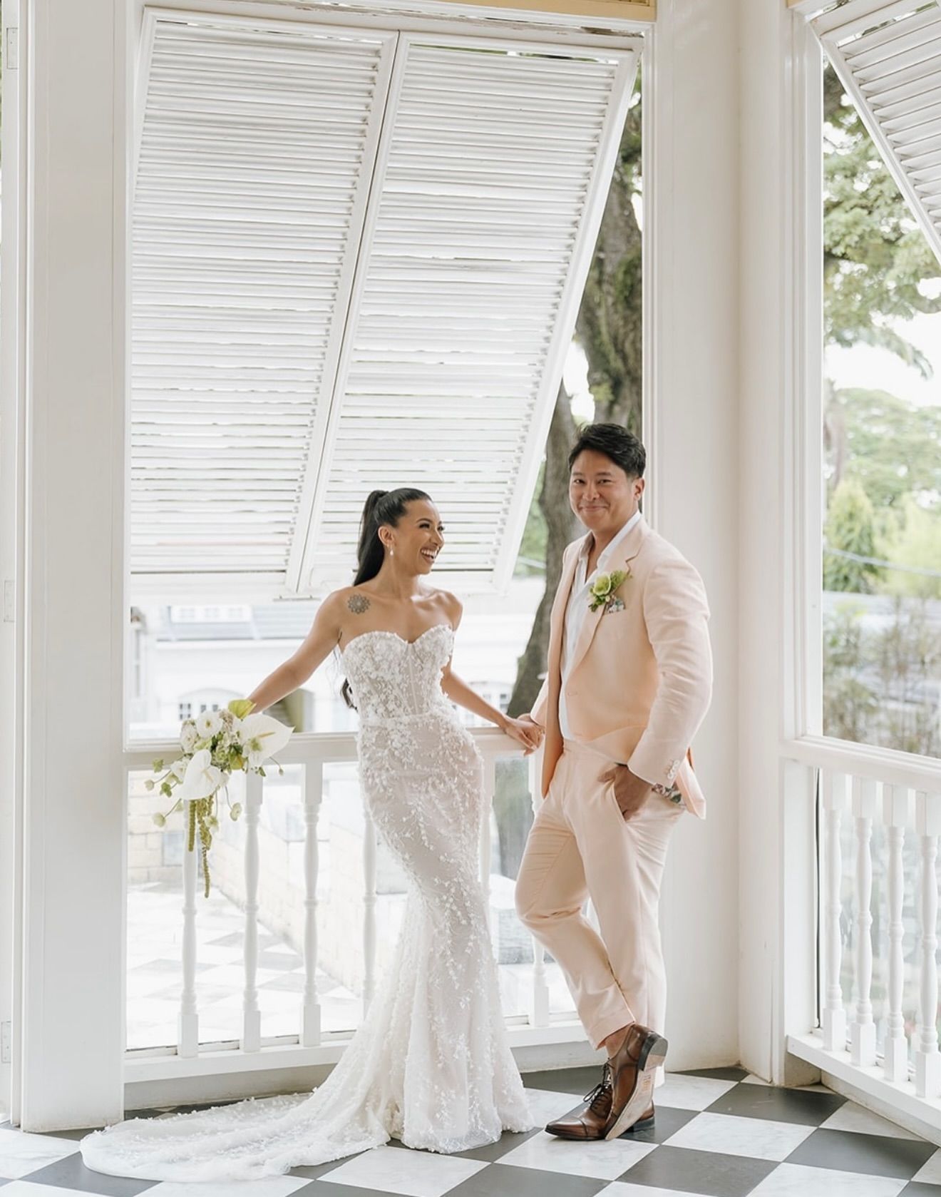 Bride and groom in wedding attire smiling, holding hands on a porch with white shutters.