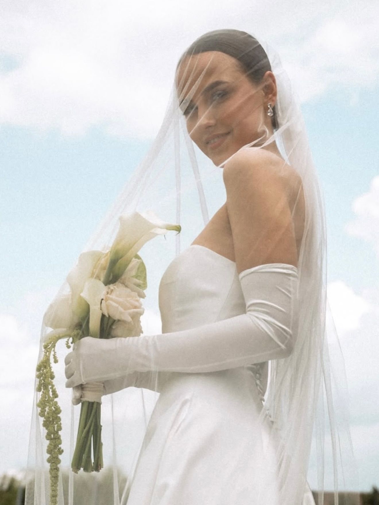 Bride in white satin dress, veil, and gloves holding bouquet, smiles, outdoor setting.