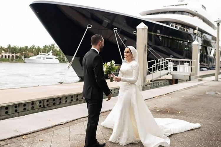 Bride and groom exchanging vows near a large yacht. Bride wears a long-sleeved white gown, groom in a black suit.
