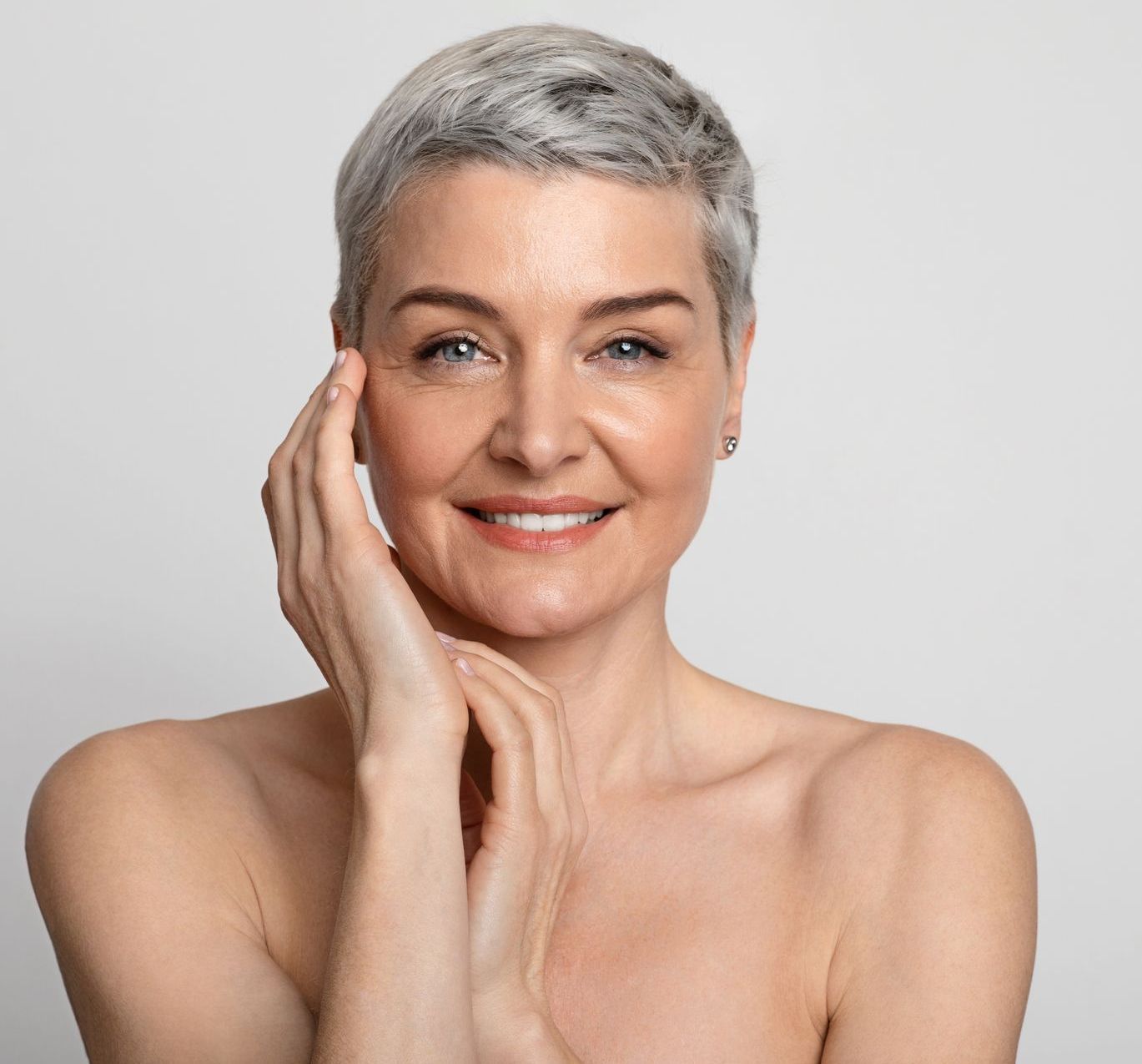 A woman with short gray hair is smiling and touching her face with her hand.