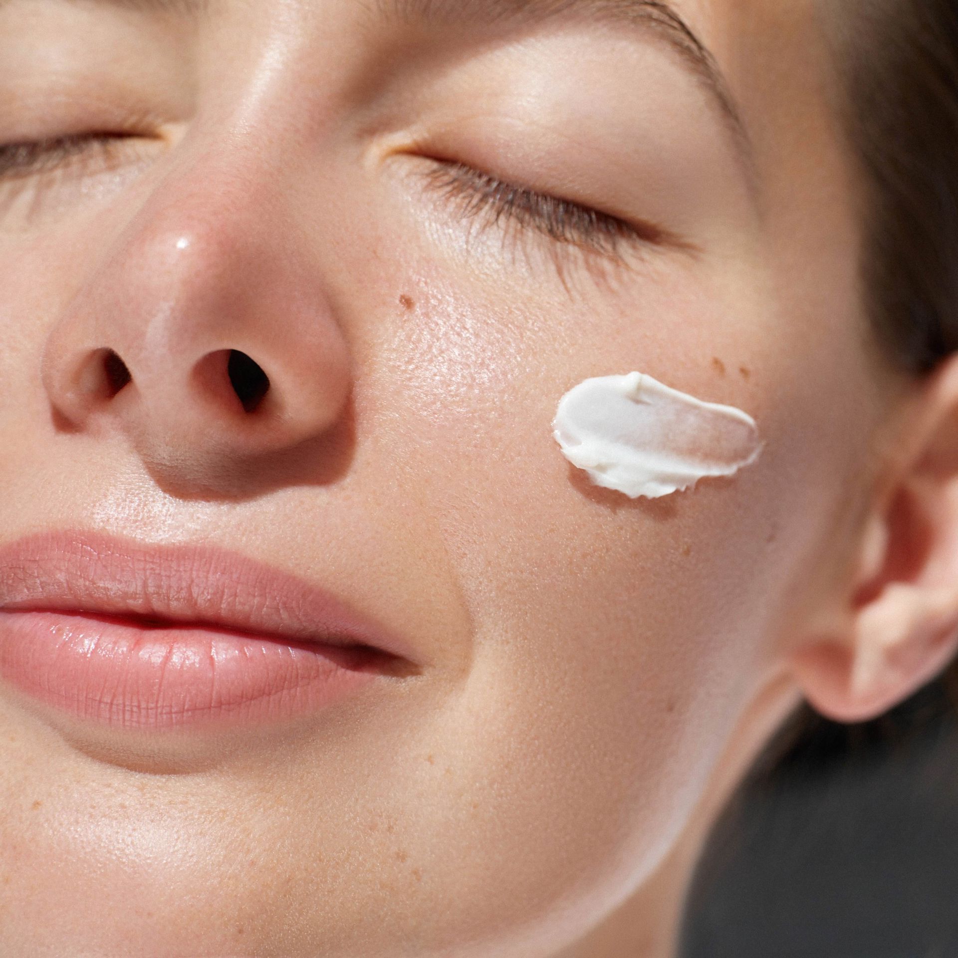 A woman is applying cream to her face with her eyes closed.